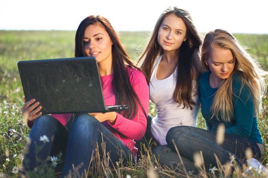 three young beautiful woman with a laptop sitting in the field on sky background 