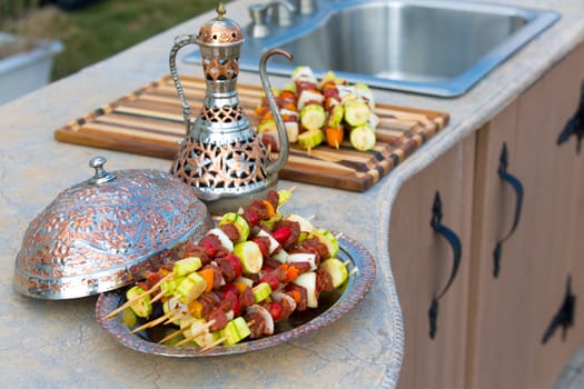 Raw skewers are waiting on the outdoor kitchen counter top. Skewers are in the copper plate  along with complimenting ornamented copper pitcher.