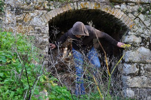 Hooded man obscured by overgrown plants under arched recess of rural ruin.