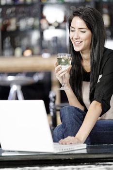 Beautiful young woman chatting with friends on her laptop while enjoying a glass of wine in a bar