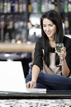 Beautiful young woman chatting with friends on her laptop while enjoying a glass of wine in a bar