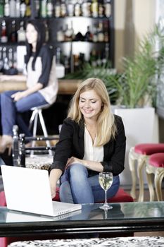 Beautiful young woman chatting with friends on her laptop while enjoying a glass of wine in a bar