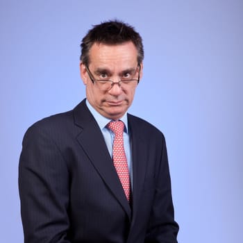 Grumpy Frowning Middle Age Business Man in Suit Looking over Glasses on Blue Background