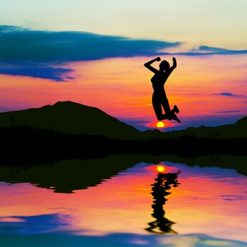 Silhouette of a happy woman jumping for joy at sunset. Water reflection