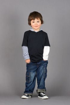 Cute and hip little preschooler boy with big brown eyes smiling with hands in his jeans pockets over grey background.