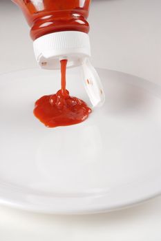 Bottle of ketchup pouring out onto a white plate.
