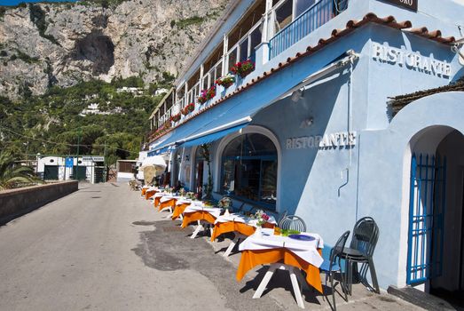 Brightly coloured tables outside blue cafe, under craggy mountains, Capri Italy