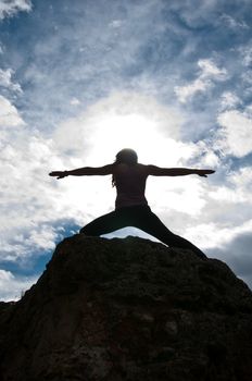 Silhouette of a young attractive girl doing the Warriors pose from yoga on top of a rock with the sky and clouds behind her.