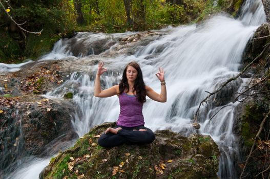 Young attractive girl sitting on a rock and meditating in a forest with a stream cascading down behind her. The water is slowed and blurred to give a dreamy looking effect.
