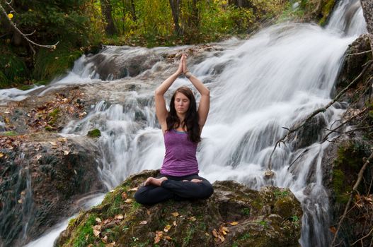 Young attractive girl sitting on a rock and meditating in a forest with a stream cascading down behind her. The water is slowed and blurred to give a dreamy looking effect.