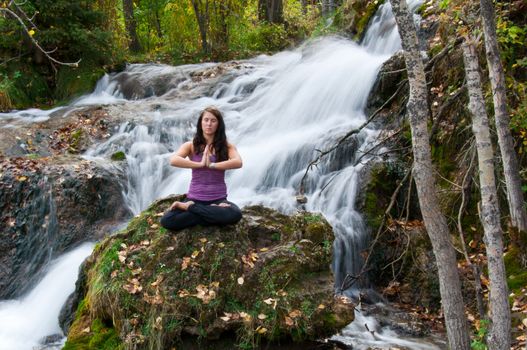 Young attractive girl sitting on a rock and meditating in a forest with a stream cascading down behind her. The water is slowed and blurred to give a dreamy looking effect.