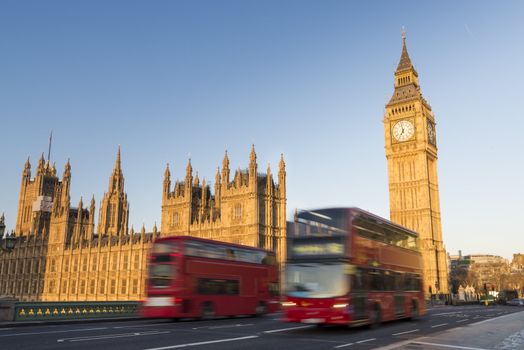 North face of Big Ben with houses of parliament, in London, UK, and motion blurred red double decker buses passing by.