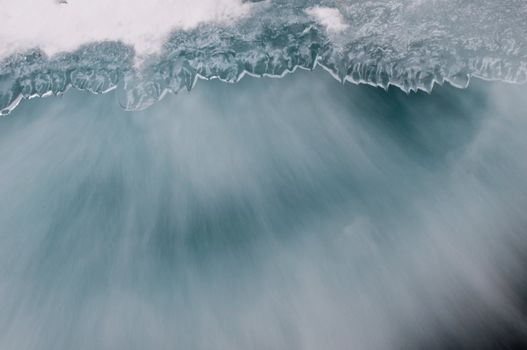 close up of the bottom of a waterfall showing movement of the water around the edge of ice through long exposure blurring creating a wintry background