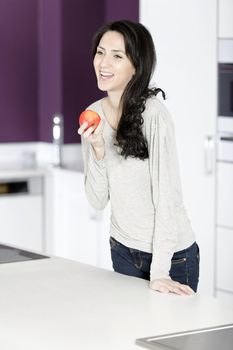 Beautiful young woman eating an apple in her white kitchen relaxing