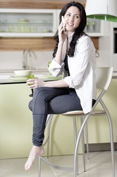 Beautiful young woman chatting on her mobile phone in her kitchen