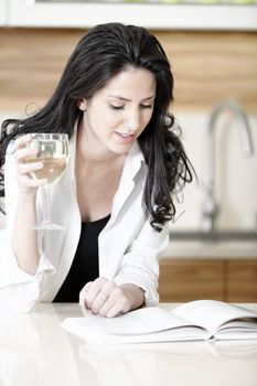 Attractive young woman reading a recipe book while enjoying a glass of wine in the kitchen