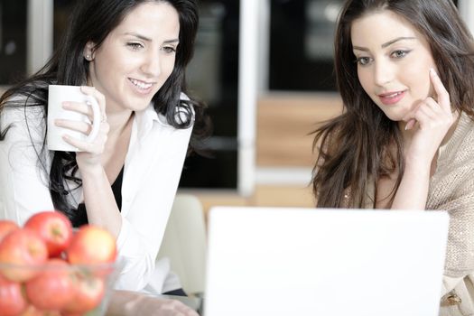 Two friends in a kitchen catching up and having fun on a laptop computer.