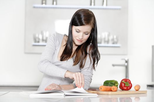 Beautiful young woman reading from a cookery book in her kitchen.