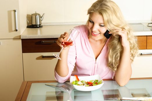 beautiful blond girl at home in the kitchen eating salad and taking on the cellphone