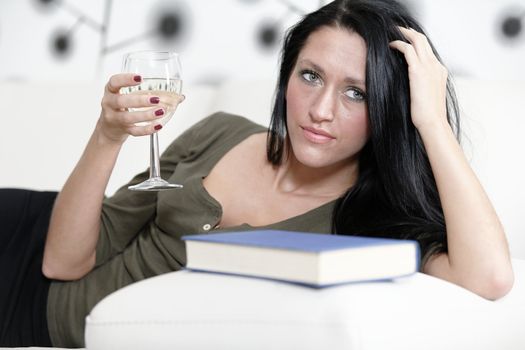 Attractive young woman lying on her sofa enjoying a glass of wine and a good book.