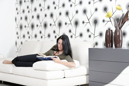 Attractive young woman lying on her sofa enjoying a glass of wine and a good book.