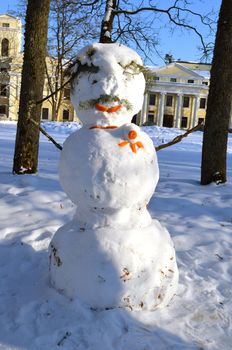 Snowman made of snow decorated with orange bark and spruce branch. Beautiful park view and old palace on backgroun..