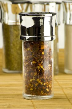 red chili flakes in a glass jar on different spices background over wooden mat