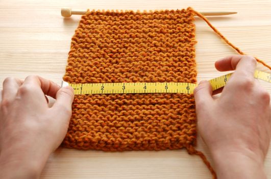 Two hands hold a tape measure across a piece of knitting on a wooden table, measuring in inches