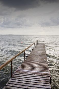 Wooden bridge on wavy lake. Red buoy in the water. Cloudy sky before storm.