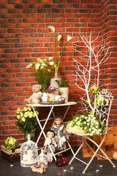 Some porcelain dolls and teddy bears on a table against a wall from a red brick