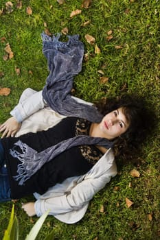 Beautiful young girl lies down on the green grass with fallen leaves.