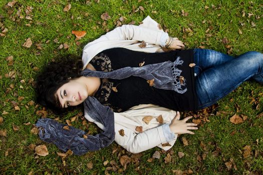 Beautiful young girl lies down on the green grass with fallen leaves.