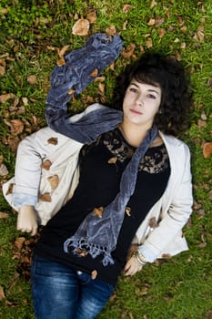 Beautiful young girl lies down on the green grass with fallen leaves.