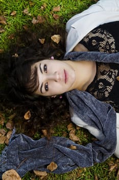 Beautiful young girl lies down on the green grass with fallen leaves.