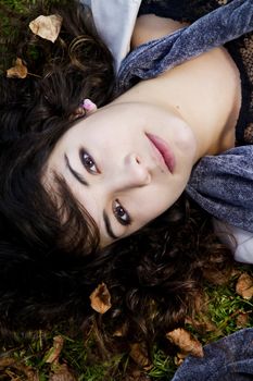 Beautiful young girl lies down on the green grass with fallen leaves.