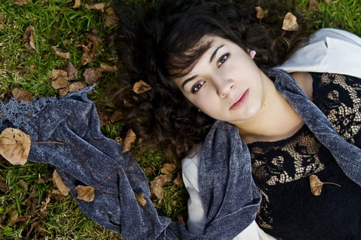 Beautiful young girl lies down on the green grass with fallen leaves.