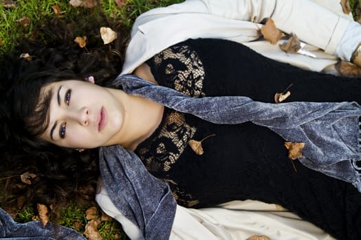 Beautiful young girl lies down on the green grass with fallen leaves.