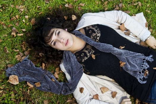 Beautiful young girl lies down on the green grass with fallen leaves.