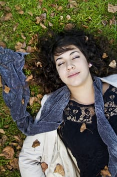 Beautiful young girl lies down on the green grass with fallen leaves.