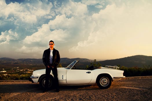 View of a young male with a jacket next to his white convertible car.