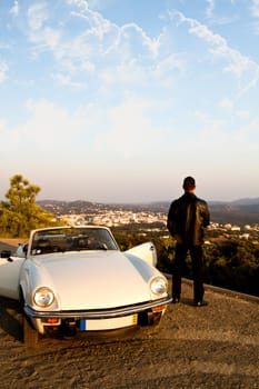 View of a young male with a jacket next to his white convertible car.