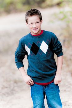 A portrait of a cute young boy wearing a sweater and jeans outdoors at a park.