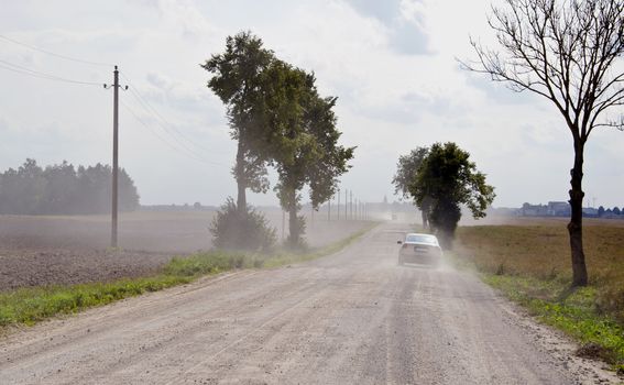 Dusty rural gravel road between farm fields and the machine going.