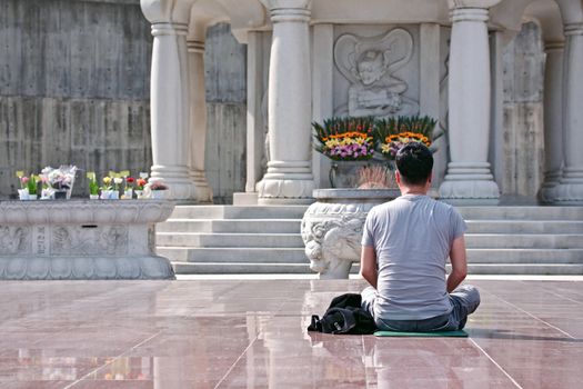 The back of a young man monk dressed in T-shirt sitting praying in front of Buddha statue