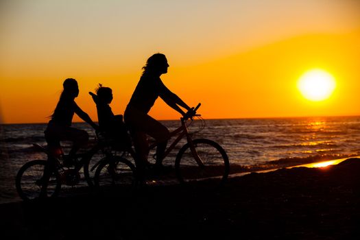 Mother and her kids on the bicycle silhouettes on beach at sunset