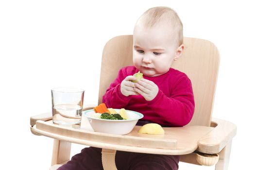 young child in red shirt eating vegetables in wooden high chair.