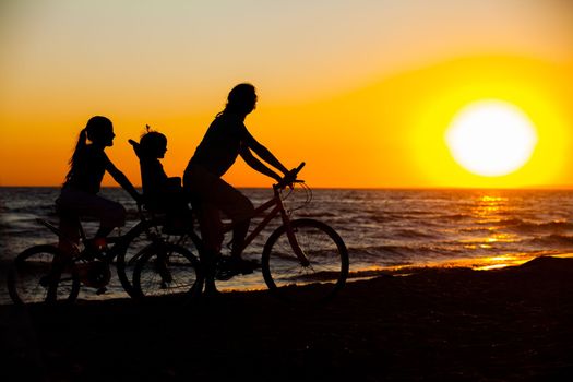 Mother and her kids on the bicycle silhouettes on beach at sunset