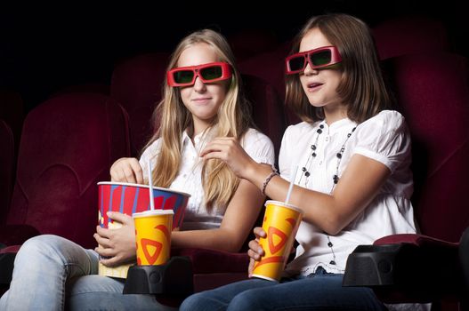 two girls look three-dimensional cinema, sitting in the glasses, eat popcorn, drink drink