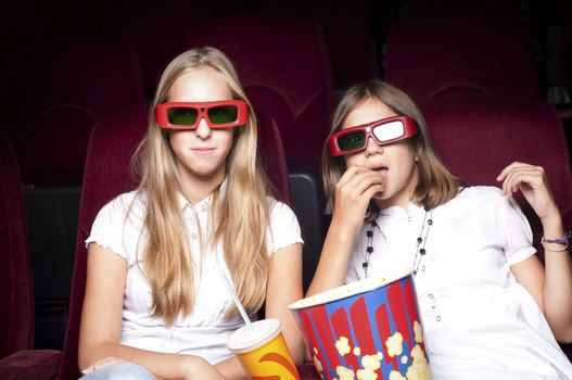 two girls look three-dimensional cinema, sitting in the glasses, eat popcorn, drink drink