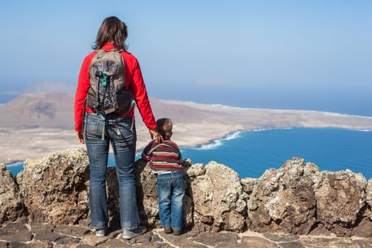Back view young mother with backpack and son standing on cliff's edge and looking to a island.